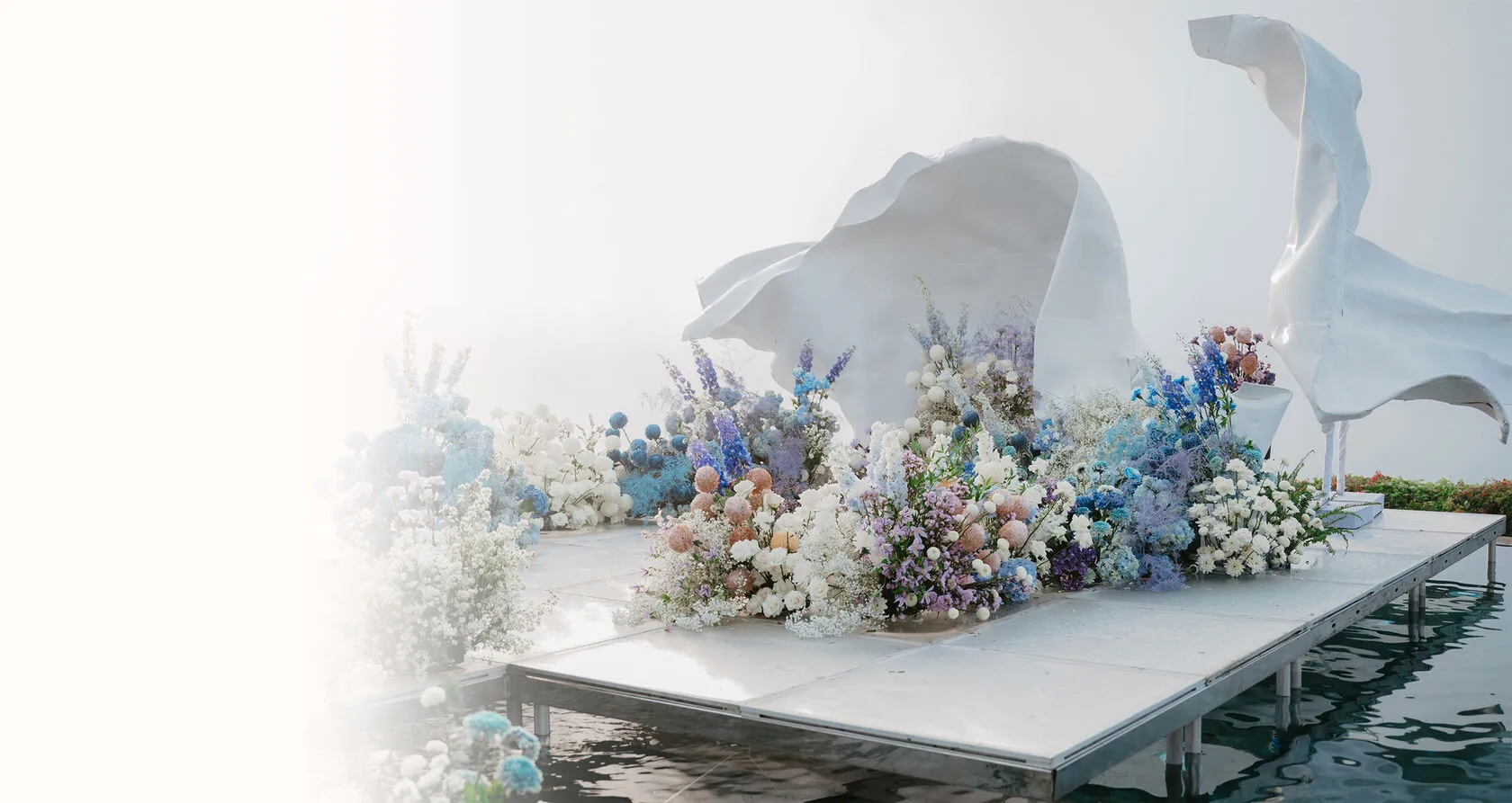 Large pastel floral installation with white sculptural drapery on a waterside stage
